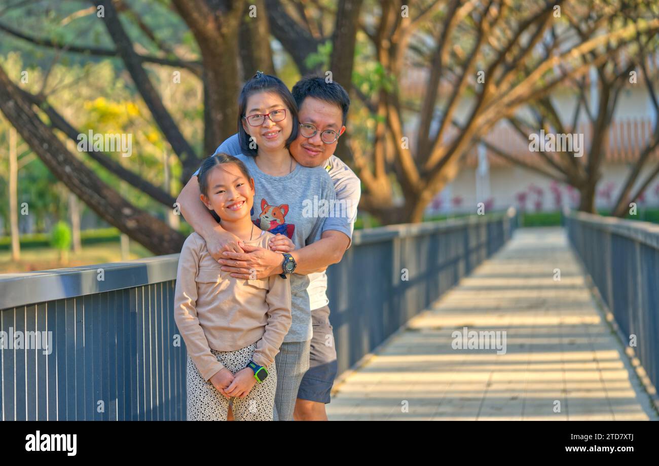 Famille asiatique heureuse en vacances dans une station balnéaire, père, mère et petite fille, debout sur un pont sous grand arbre dans le jardin, vue en perspective, n Banque D'Images