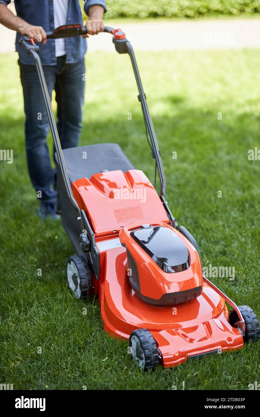 Travailleur anonyme masculin tondant la pelouse de jardin avec un nouveau coupe-herbe sans fil. Vue du dessus de la tondeuse à gazon orange brillant moderne, actionnée par un bricoleur, on Banque D'Images Travailleur anonyme masculin tondant la pelouse de jardin avec un nouveau coupe-herbe sans fil. Vue du dessus de la tondeuse à gazon orange brillant moderne, actionnée par un bricoleur, on Banque D'Images