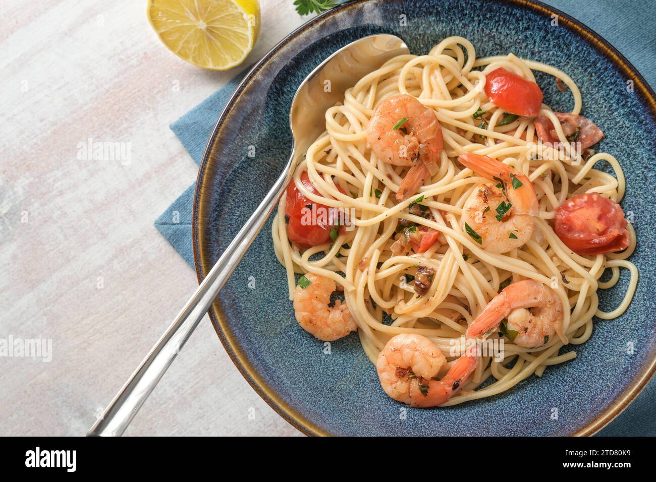 Spaghetti aux crevettes, tomates et herbes en sauce citron dans une assiette bleue avec cuillère sur une table en bois blanc, fruits de mer méditerranéens maison, copie spa Banque D'Images