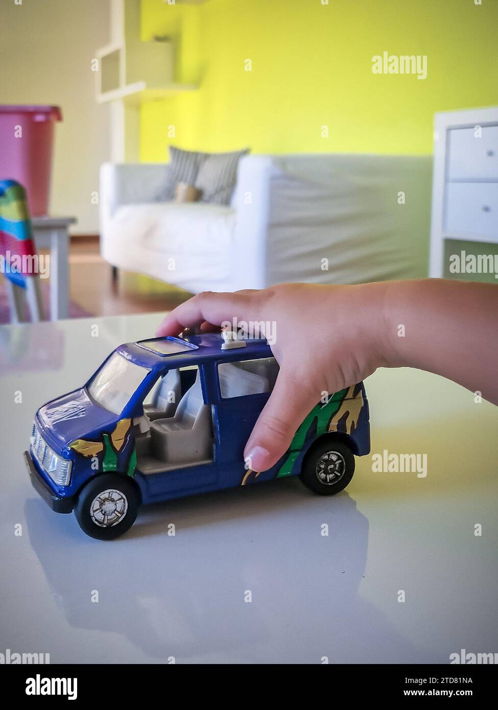 Gros plan de la petite main d'un enfant saisissant une voiture jouet bleue vibrante sur une salle de jeux colorée, encapsulant la joie et l'innocence du jeu de l'enfance Banque D'Images