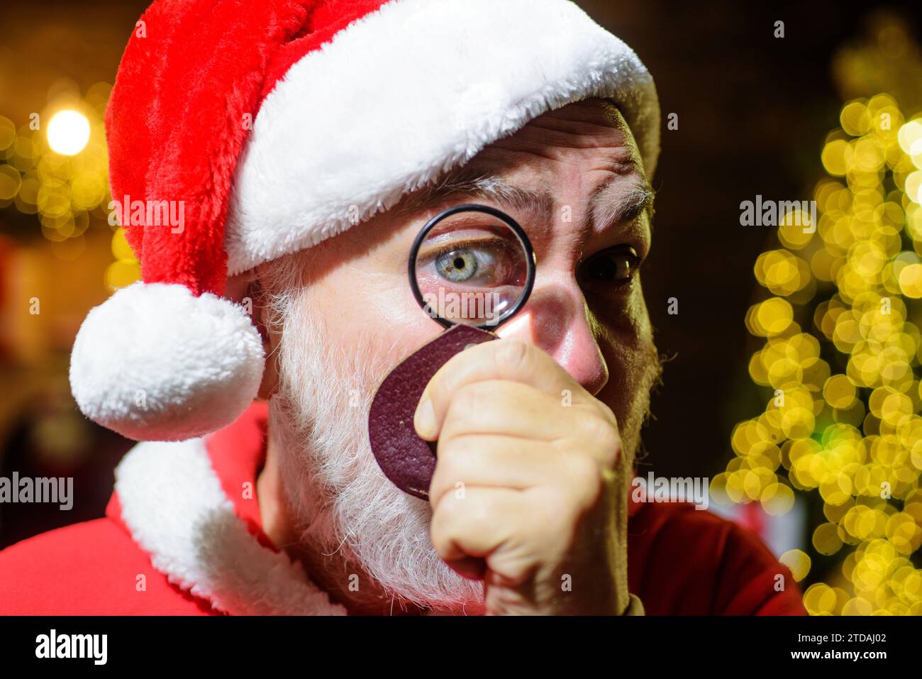 Christmastime. Père Noël regardant à travers la loupe. Gros plan portrait homme barbu dans les vêtements du Père Noël avec lentille loupe. Joyeux Noël Banque D'Images