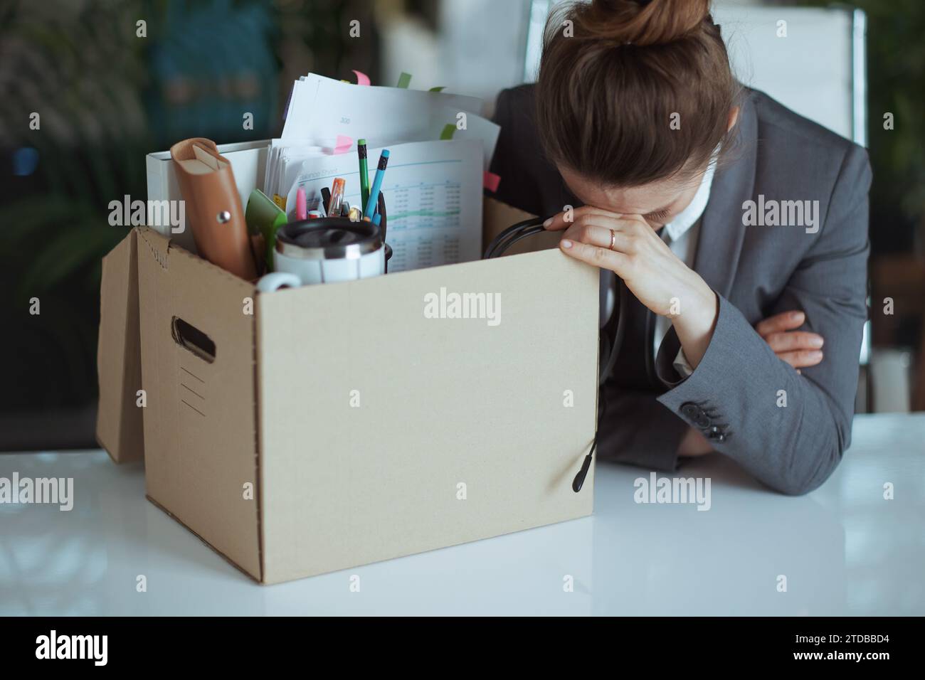 Nouveau travail. femme travailleuse moderne malheureuse dans le bureau vert moderne en costume gris d'affaires avec des effets personnels dans la boîte en carton. Banque D'Images