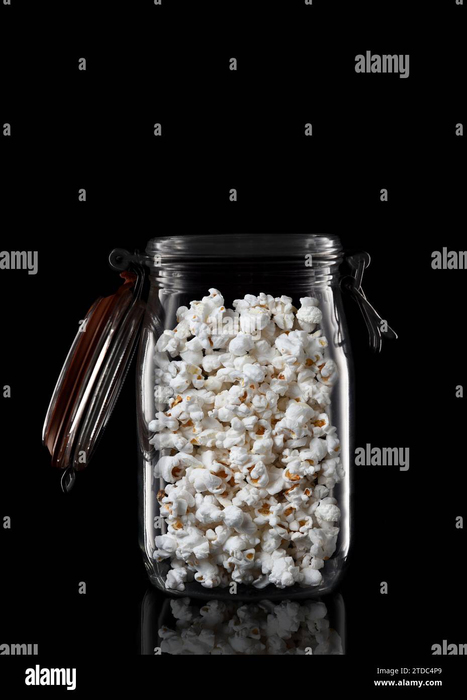 Un pot de stockage ou de mise en conserve en verre rempli de pop-corn isolé sur noir avec reflet, avec couvercle ouvert. Banque D'Images