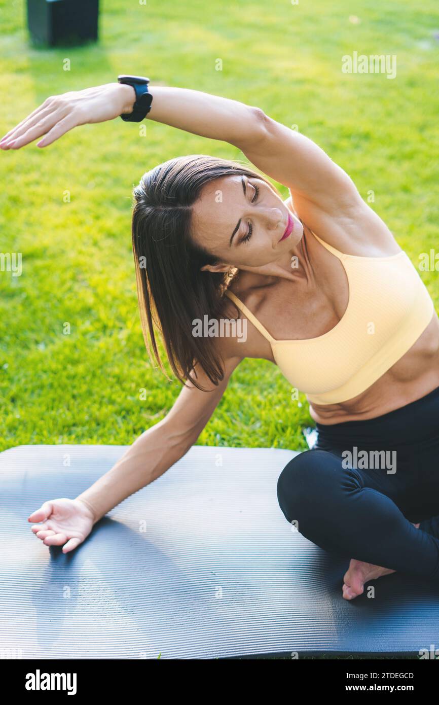 Femme caucasienne sportive attrayante assise sur un tapis de yoga, faisant des exercices d'étirement au parc urbain. Femme positive travaillant sur la flexibilité des muscles Banque D'Images