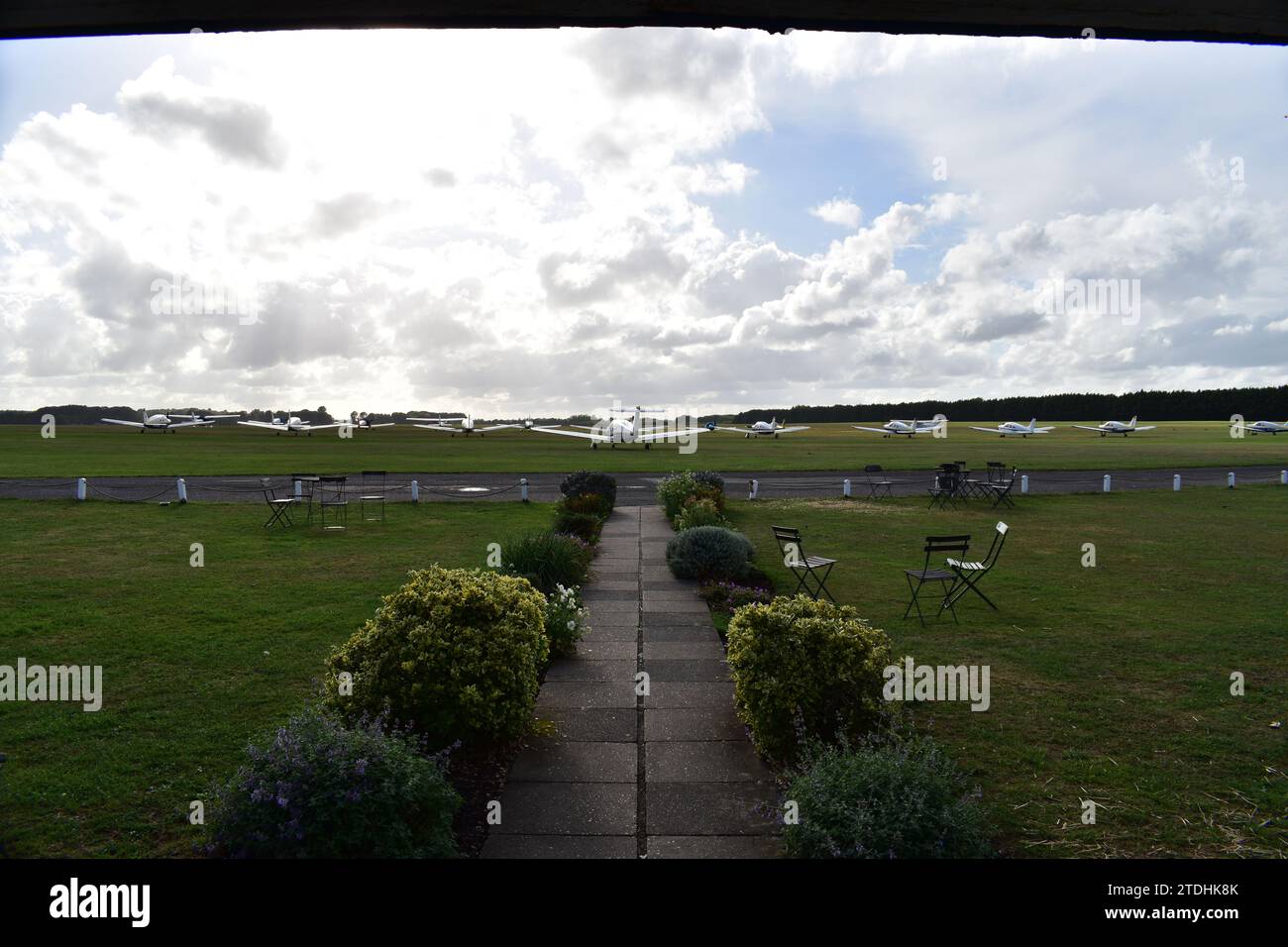 Un chemin de tuiles de pierre décoré de buissons luxuriants à côté d'un champ d'herbe avec des chaises menant à un champ d'herbe avec plusieurs petits avions monomoteurs Banque D'Images