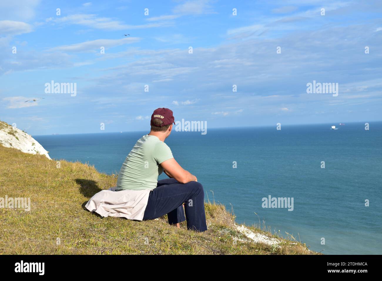 Jeune homme portant un pantalon bleu foncé, un t-shirt vert clair et une casquette bordeaux assis sur le bord des falaises blanches de Douvres surplombant la mer Banque D'Images