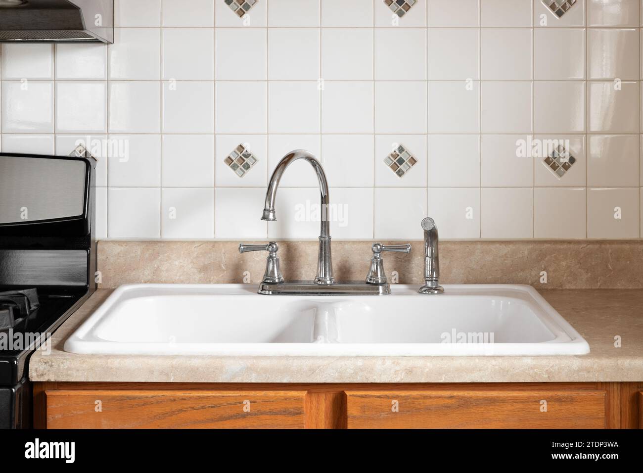 Un vieux détail de robinet de cuisine avec armoires en bois, évier blanc, robinet chromé, et un dosseret de carrelage carré. Banque D'Images