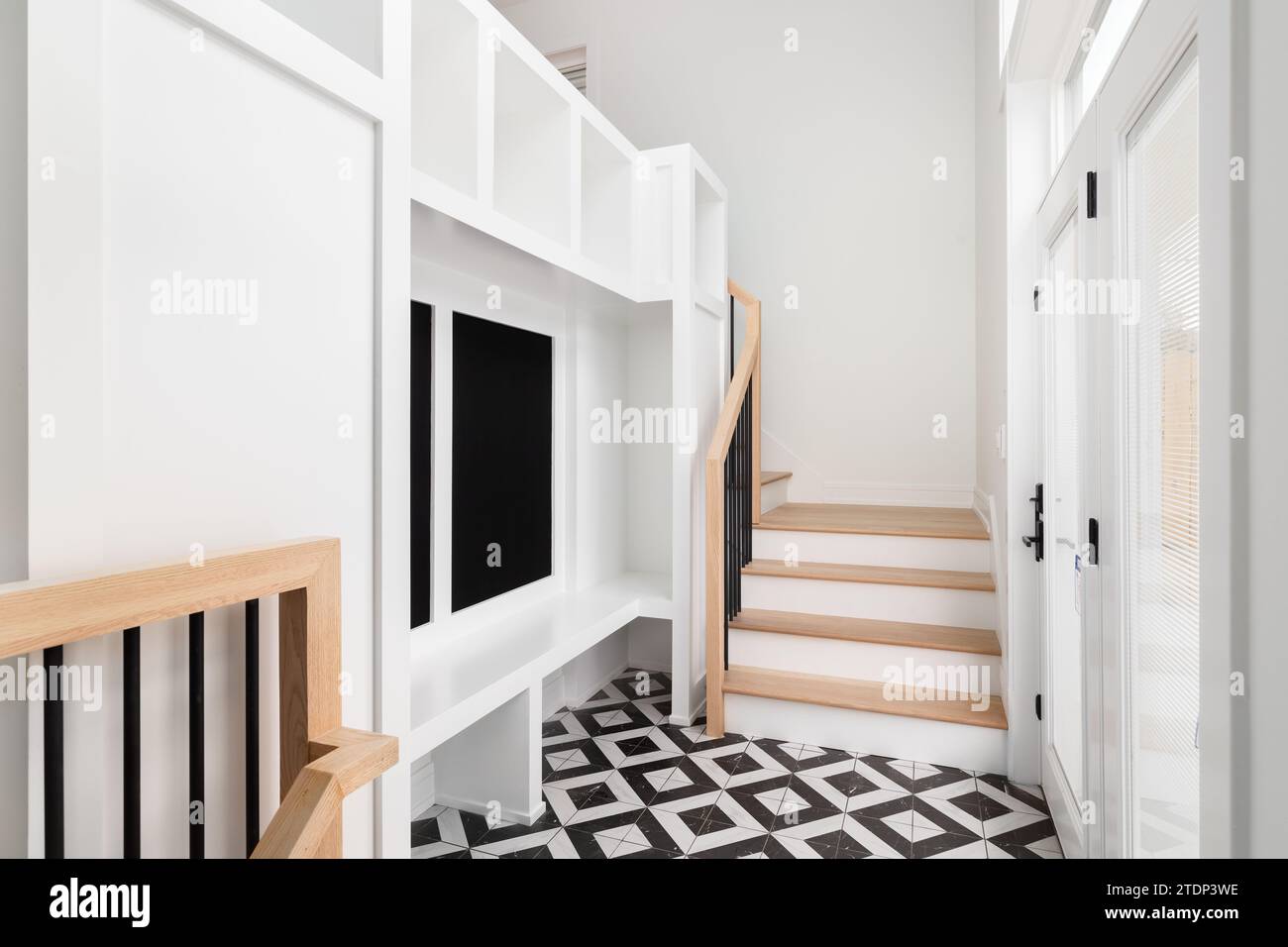 Un grand foyer avec une unité de rangement blanche, banc blanc, escalier et balustrade en chêne blanc, et un sol carrelé noir et blanc. Banque D'Images