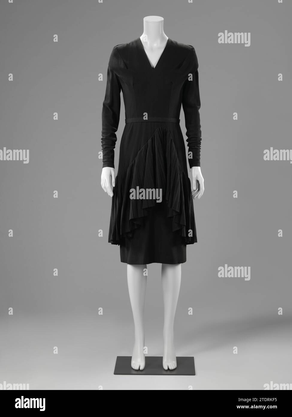 Robe avec flonce plissé, Catharina Kruysveldt-de Mare, c. 1948 - c. 1954 Cocktailjapon de laine noire avec Volan plissé large sur la jupe. Modèle : col en V et manches longues et ajustées avec fermeture éclair en métal au poignet. Une bande lâche sur les épaules, le long du cou jusqu'au Coupenaden, qui donne l'idée d'une ligne d'épaule élargie. Jupe étroite sur laquelle une seconde jupe plus large avec une bande plissée de 53 cm de long, qui monte à 4,5 cm sous la ceinture au milieu. Fermeture au milieu derrière une fermeture éclair métallique. Unsurbed. licencié : AmsterdamParis entier : laine. Fermeture : cocktail en métal Banque D'Images