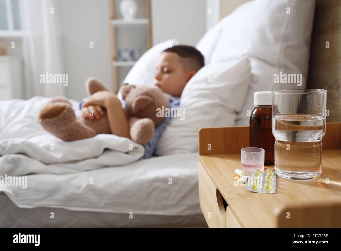 Pilules avec du sirop contre la toux et de l'eau sur la table du petit garçon malade dans la chambre, gros plan Banque D'Images