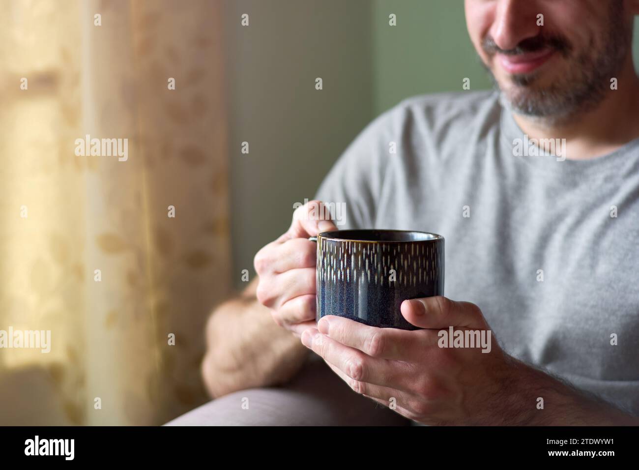 Gros plan d'un jeune homme caucasien souriant et tenant une tasse de thé. Homme joyeux appréciant le thé chaud dans son salon à la maison, se relaxant pendant sa pause. C Banque D'Images