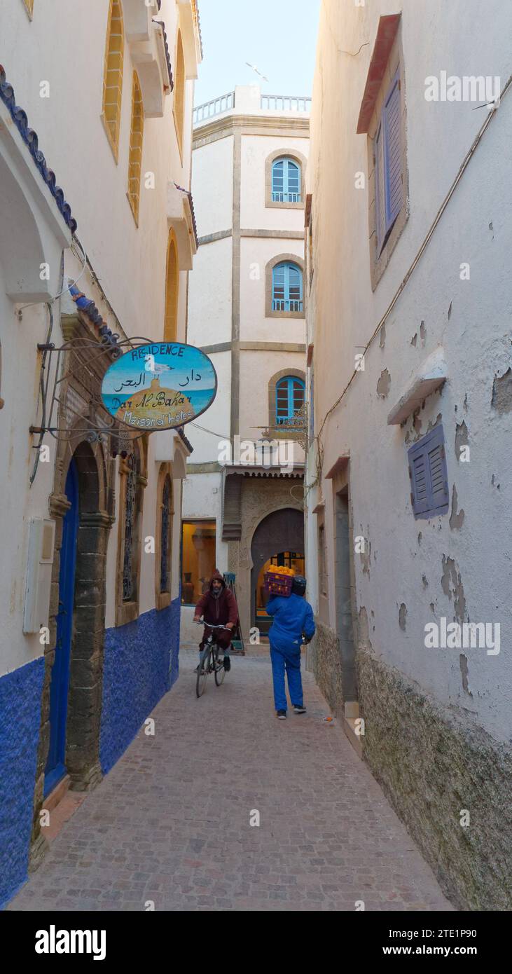 Étroite rue bleue et blanche avec un hôtel dans la médina avec un cycliste et un travailleur portant un plateau d'oranges. Essaouira, Maroc. 19 décembre 2023 Banque D'Images