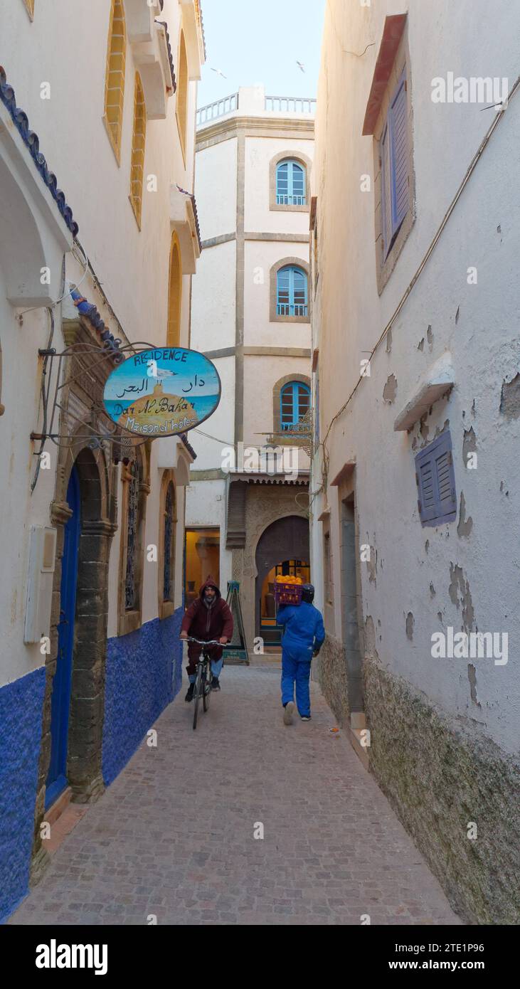 Étroite rue bleue et blanche avec un hôtel dans la médina avec un cycliste et un travailleur portant un plateau d'oranges. Essaouira, Maroc. 19 décembre 2023 Banque D'Images