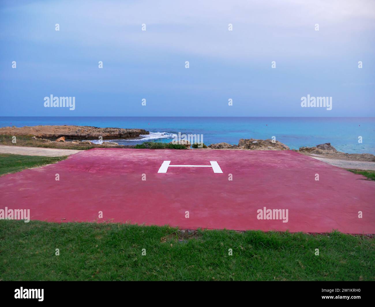Coussin d'hélicoptère rouge avec une lettre H au centre sur la rive de la mer Méditerranée avec la vue marine et pelouse verte au premier plan. Banque D'Images
