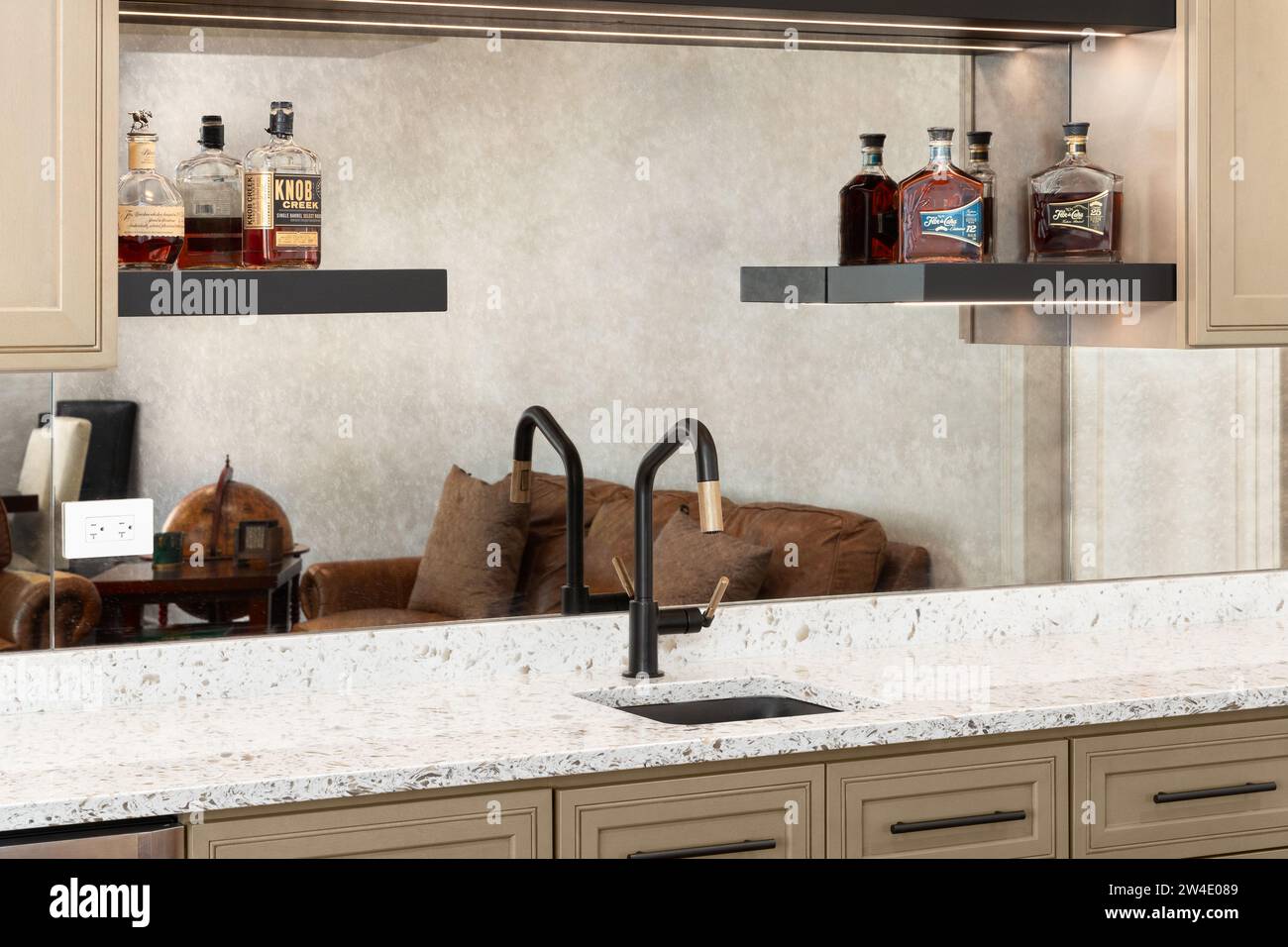 Un détail de robinet de bar humide avec des armoires brunes, un comptoir en quartz blanc, des bouteilles de whisky sur des étagères et un robinet noir et or. Banque D'Images