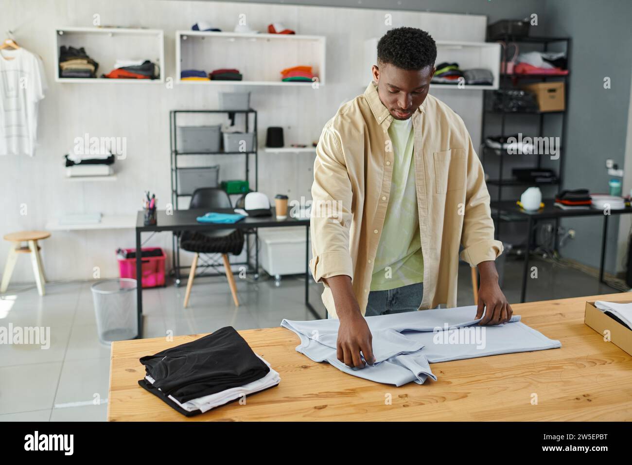 jeune et diligent homme afro-américain pliant des vêtements sur la table dans le studio d'impression, petite entreprise Banque D'Images