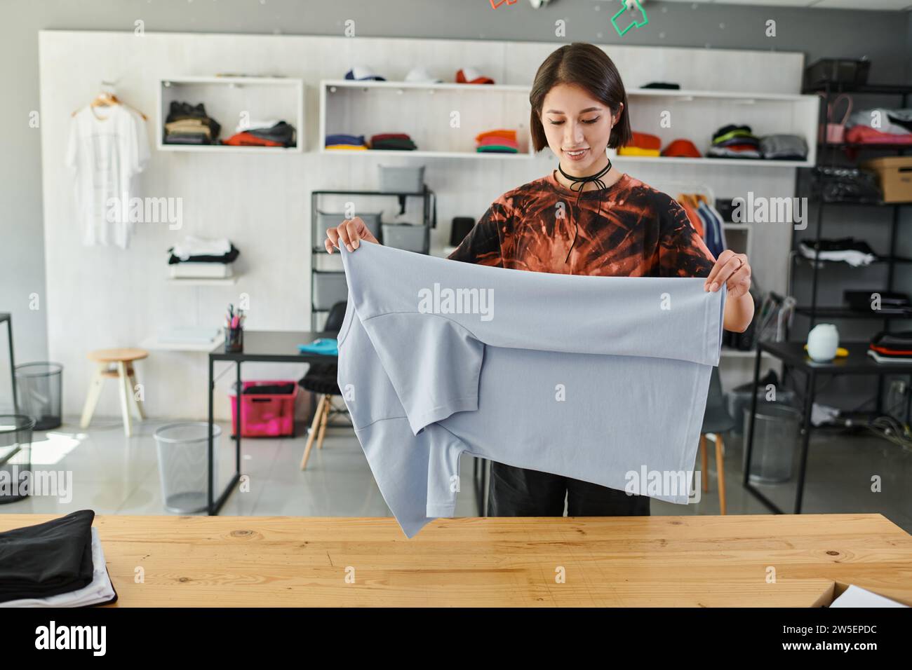 femme asiatique heureuse et qualifiée pliant les vêtements soigneusement dans le studio d'impression textile, petite entreprise Banque D'Images