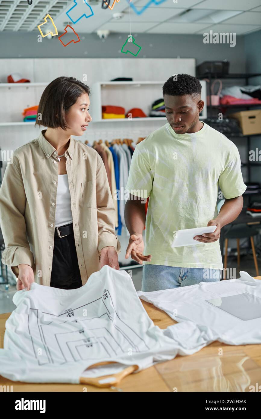 jeunes propriétaires de petites entreprises interraciales parlant près de t-shirts avec des tailles de format dans le studio d'impression Banque D'Images