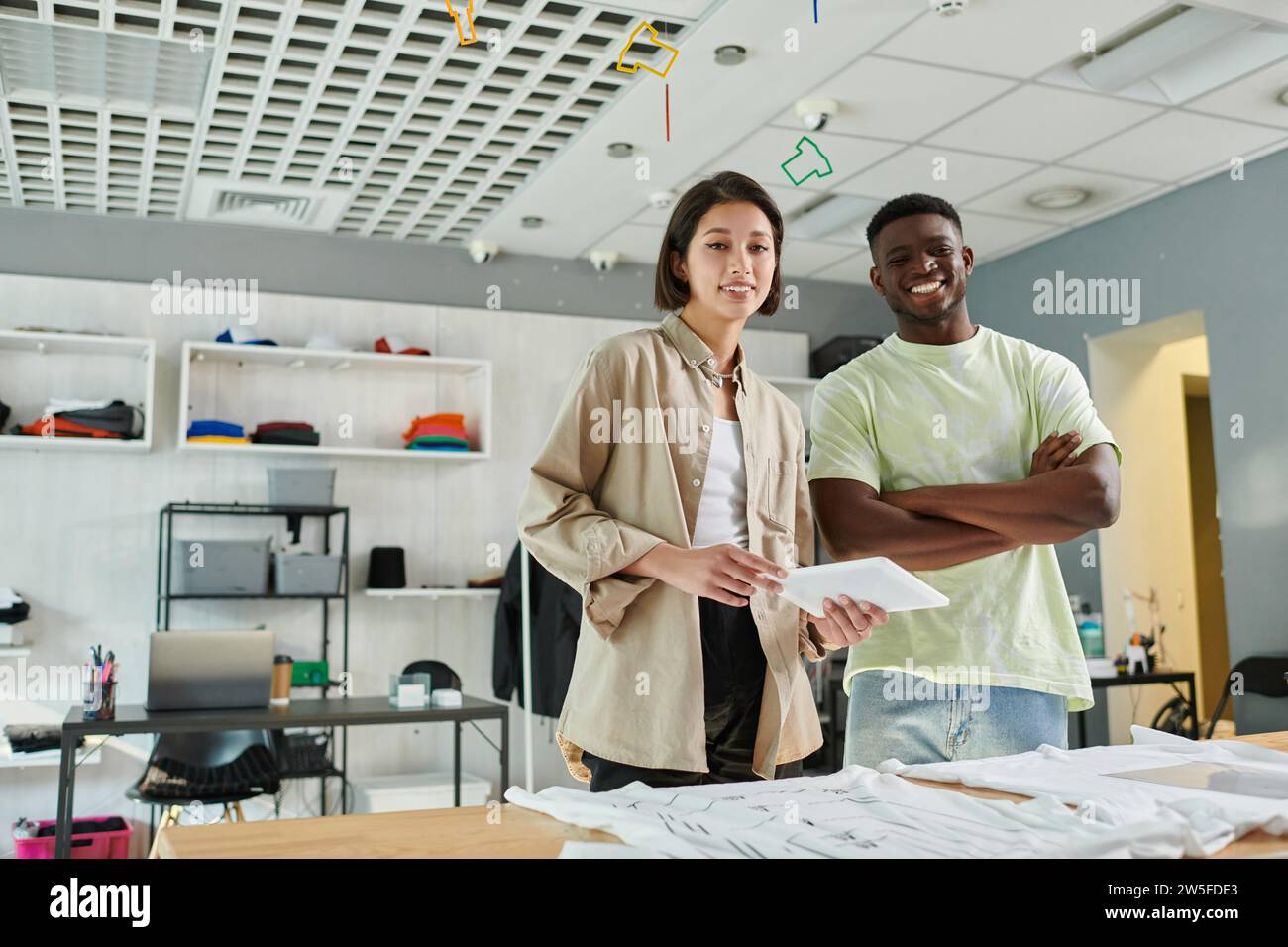 créateurs multiethniques souriant à la caméra près des vêtements dans le studio d'impression, petite entreprise Banque D'Images