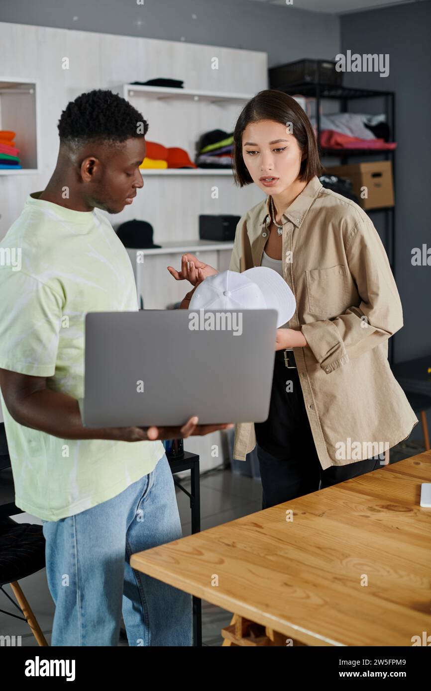 jeunes entrepreneurs interraciaux avec casquette blanche et ordinateur portable discutant de la nouvelle startup dans le studio d'impression Banque D'Images