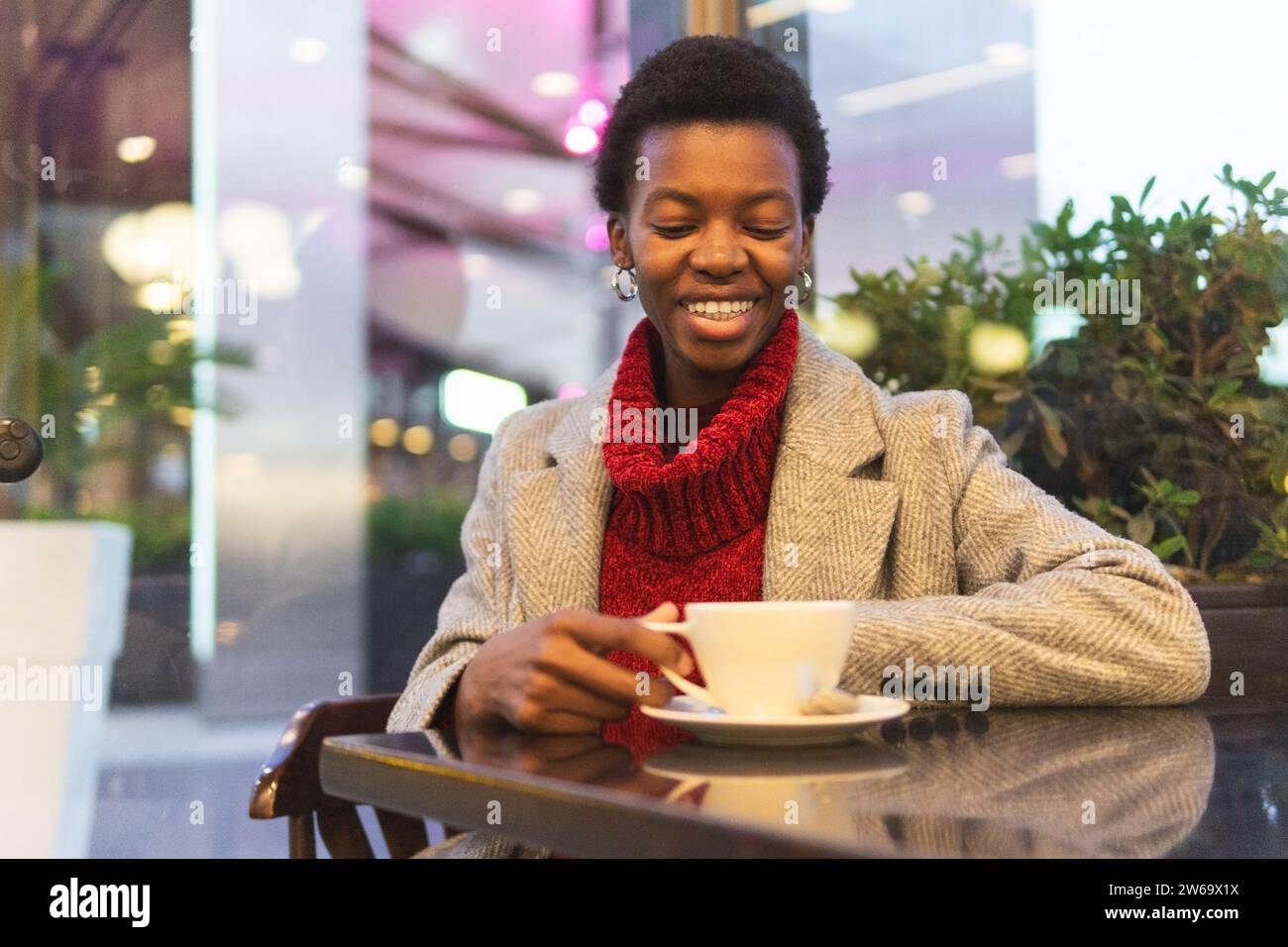 Souriante femme afro-américaine en manteau assise à table près du mur de verre buvant une boisson chaude pendant le week-end Banque D'Images
