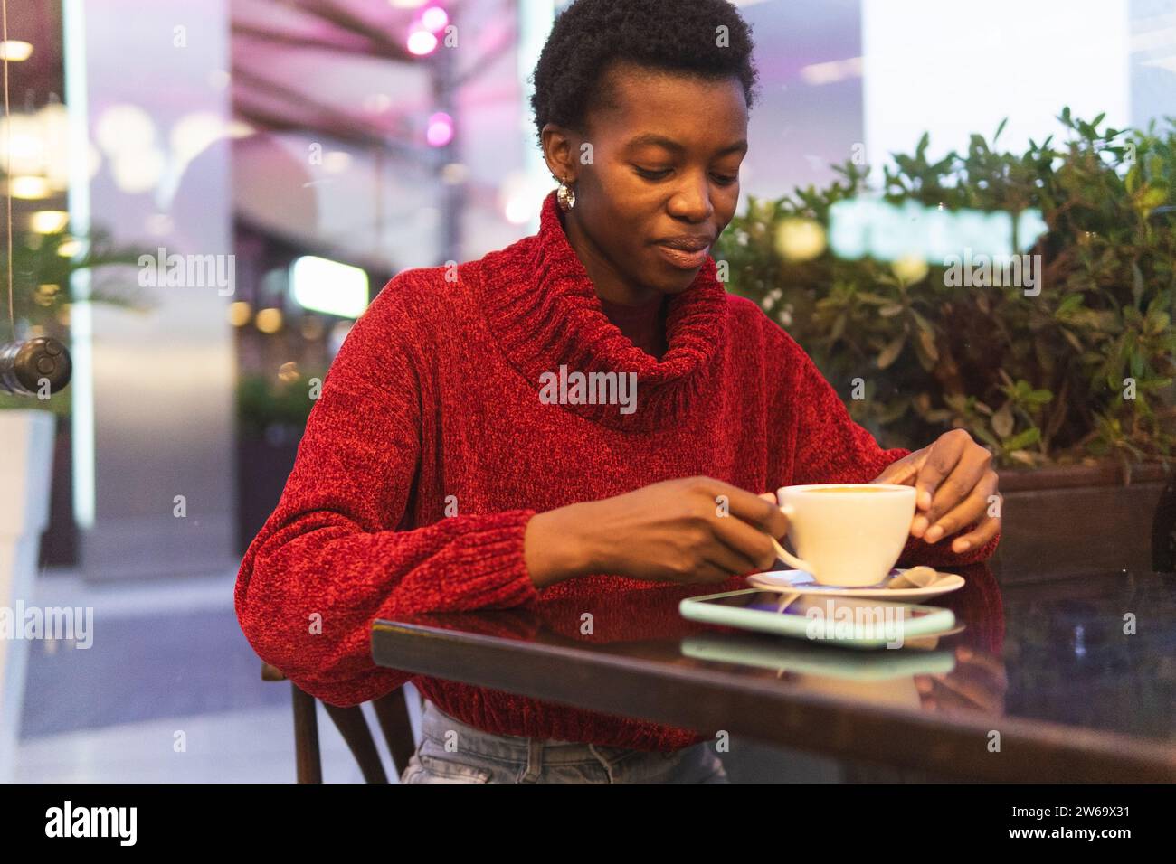 Femme afro-américaine réfléchie en manteau assise à table près du mur de verre buvant une boisson chaude pendant le week-end Banque D'Images