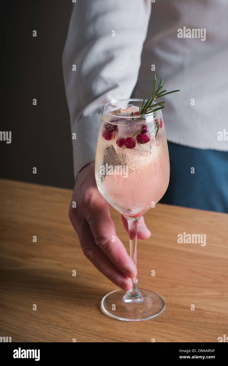 Une main présentant un cocktail d'hiver au romarin dans un verre à pied, garni de canneberges et d'une branche de romarin, sur fond neutre. Banque D'Images