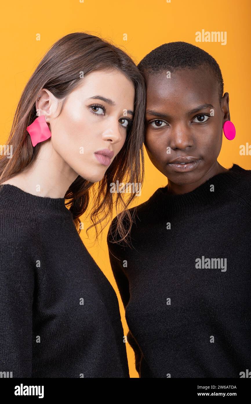 Portrait de jeunes amies multiraciales confiantes portant des boucles d'oreilles roses à la mode et des t-shirts noirs se tenant debout devant la caméra contre le dos jaune Banque D'Images Portrait de jeunes amies multiraciales confiantes portant des boucles d'oreilles roses à la mode et des t-shirts noirs se tenant debout devant la caméra contre le dos jaune Banque D'Images