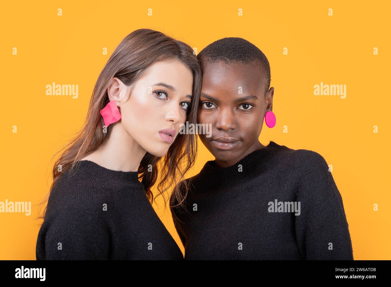 Portrait de jeunes amies multiraciales confiantes portant des boucles d'oreilles roses à la mode et des t-shirts noirs se tenant debout devant la caméra contre le dos jaune Banque D'Images Portrait de jeunes amies multiraciales confiantes portant des boucles d'oreilles roses à la mode et des t-shirts noirs se tenant debout devant la caméra contre le dos jaune Banque D'Images