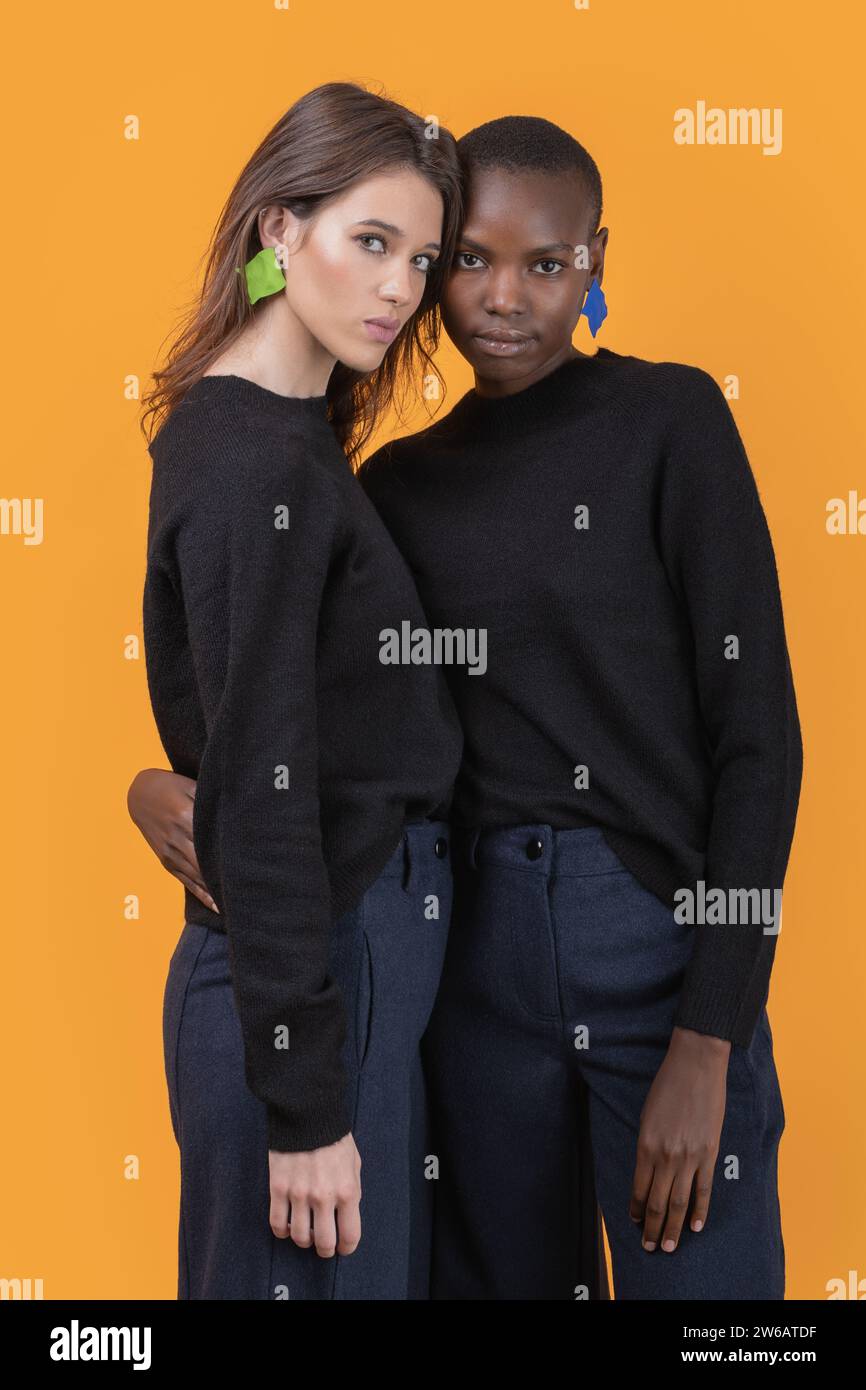 Portrait de jeunes amies multiraciales confiantes portant des boucles d'oreilles roses à la mode et des t-shirts noirs debout s'embrassant les uns les autres tout en regardant Banque D'Images Portrait de jeunes amies multiraciales confiantes portant des boucles d'oreilles roses à la mode et des t-shirts noirs debout s'embrassant les uns les autres tout en regardant Banque D'Images