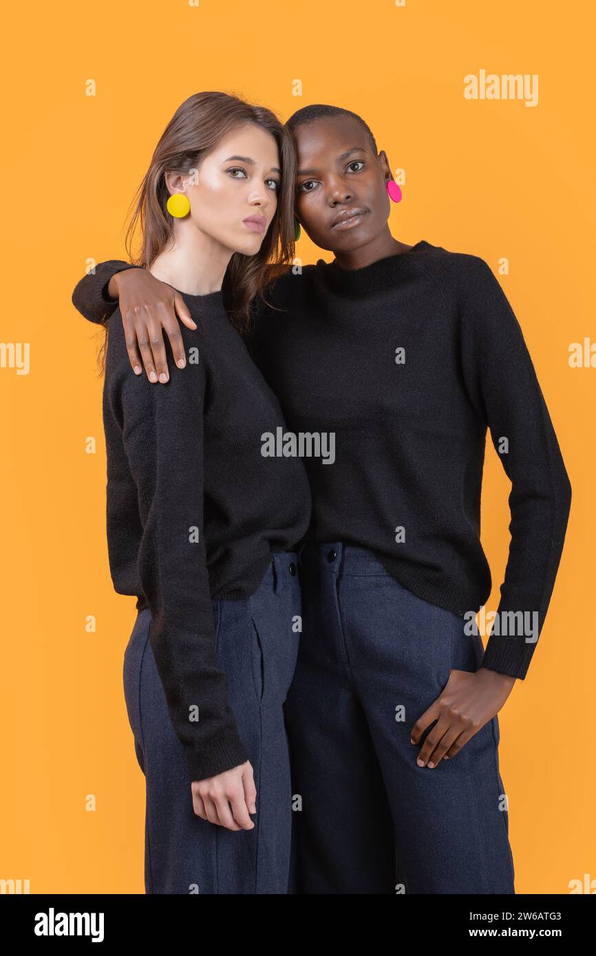 Portrait de jeunes amies multiraciales confiantes portant des boucles d'oreilles colorées à la mode et des t-shirts noirs debout s'embrassant les uns les autres tout en regardant Banque D'Images Portrait de jeunes amies multiraciales confiantes portant des boucles d'oreilles colorées à la mode et des t-shirts noirs debout s'embrassant les uns les autres tout en regardant Banque D'Images
