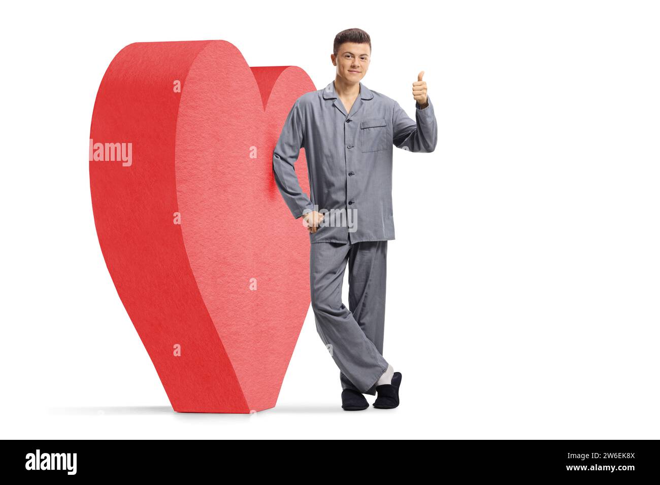 Portrait en longueur d'un gars en pyjama appuyé sur un coeur rouge et gestuant un signe de pouce vers le haut isolé sur fond blanc Banque D'Images