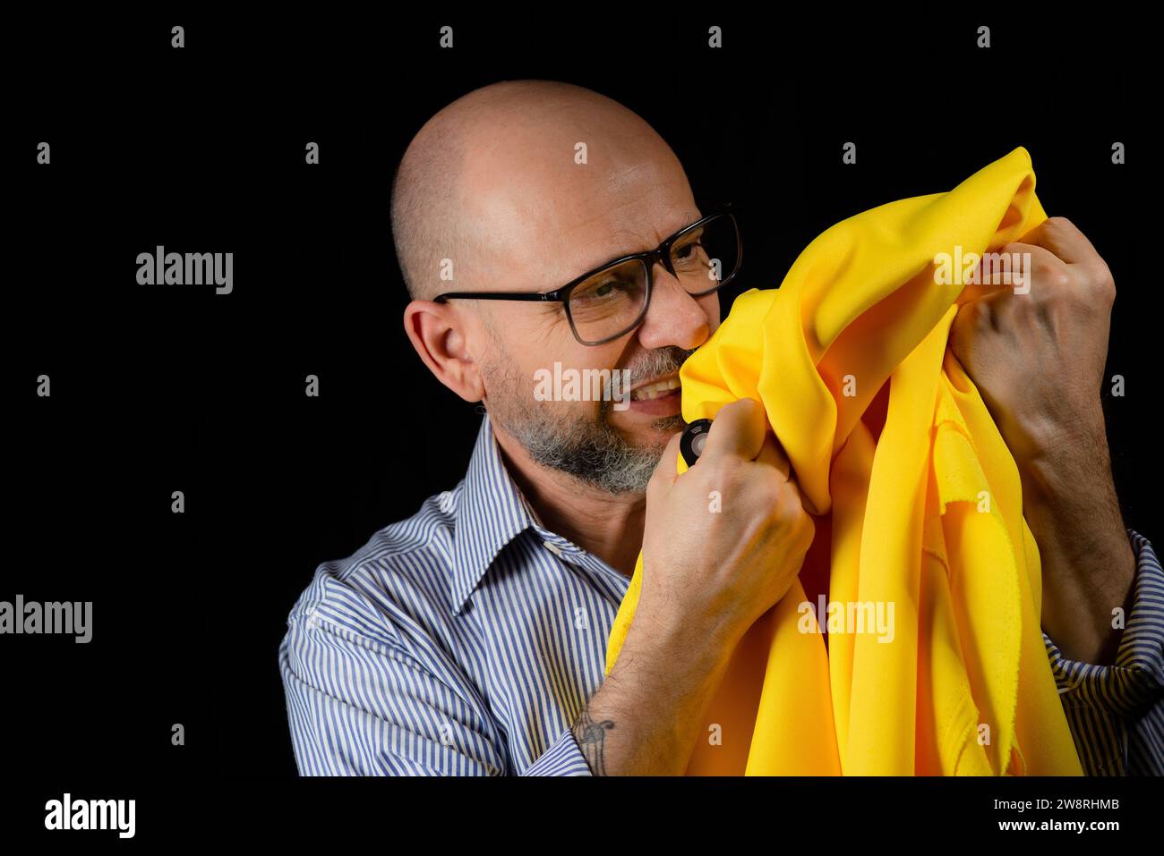 Homme chauve, barbu avec des lunettes tenant un tissu de couleur jaune au-dessus de sa tête sur fond noir. Banque D'Images