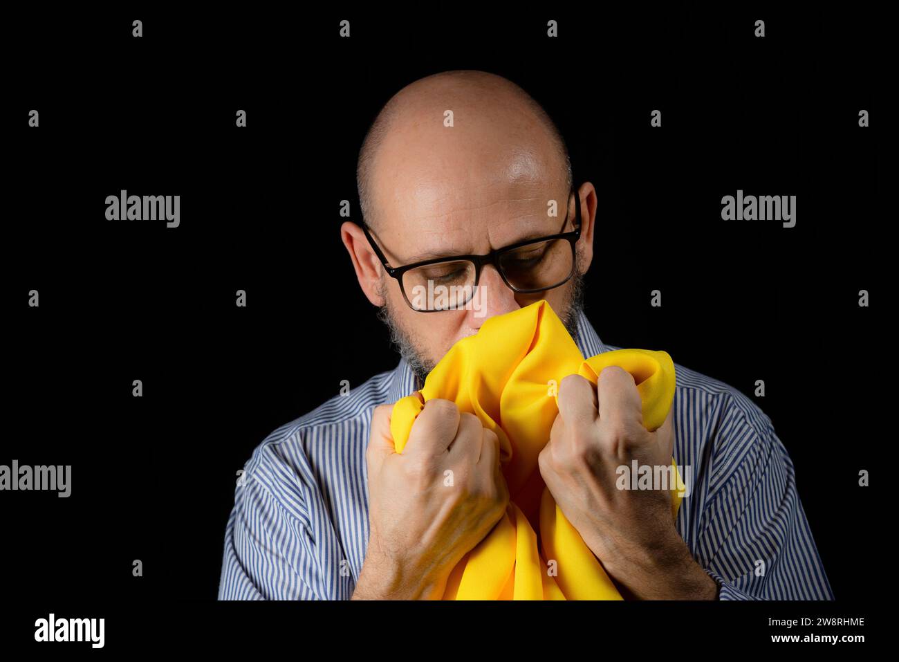 Homme chauve, barbu avec des lunettes tenant un tissu de couleur jaune au-dessus de sa tête sur fond noir. Banque D'Images