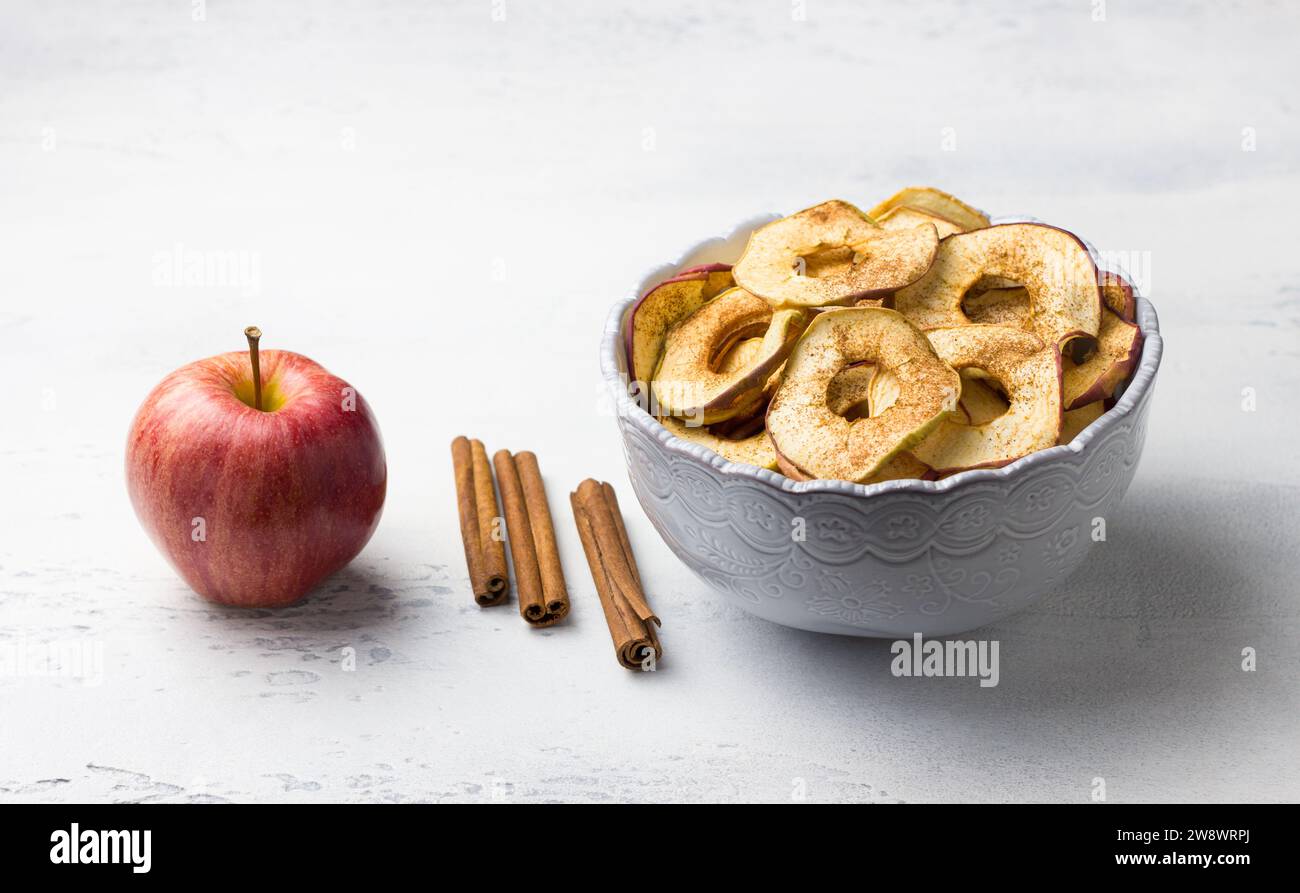 Pommes séchées maison à la cannelle, chips de pomme dans un bol avec des bâtons de cannelle sur un fond gris clair. Délicieux en-cas sain Banque D'Images