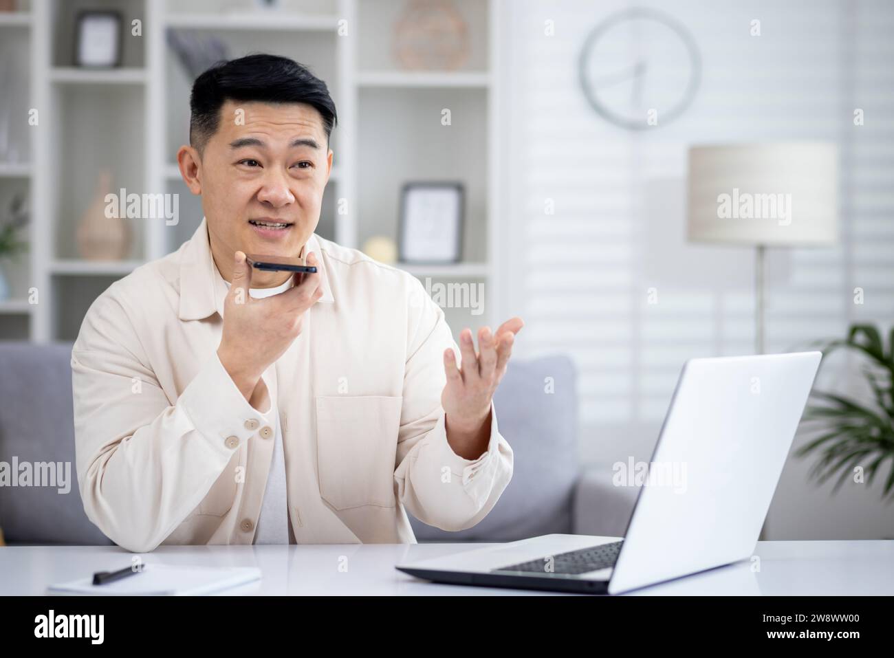 Jeune homme asiatique assis à la maison à table et travaillant à distance sur ordinateur portable, parlant sur haut-parleur, en utilisant la recherche vocale. Banque D'Images