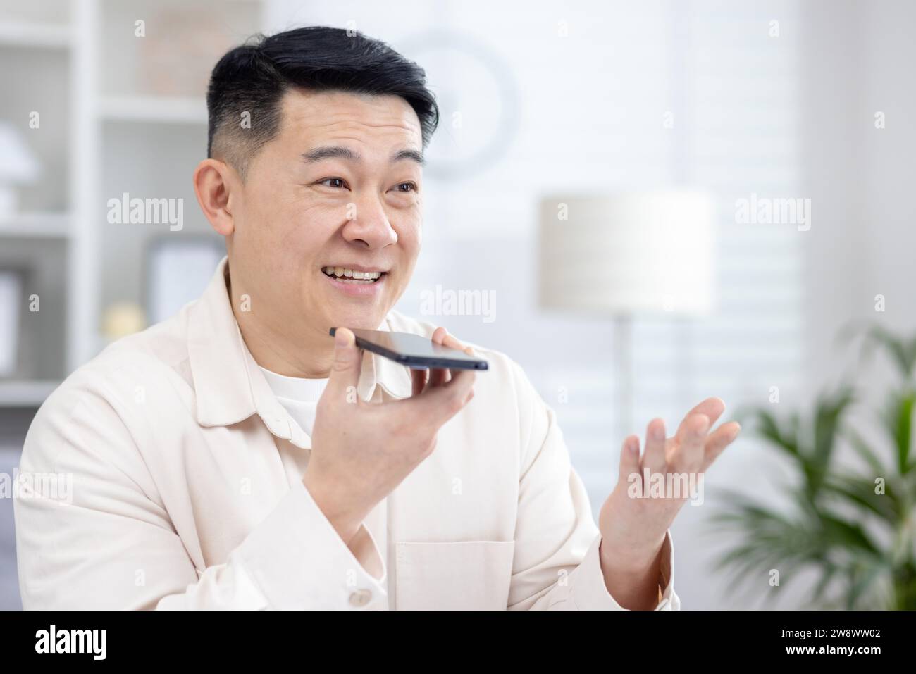 Photo en gros plan d'un jeune homme asiatique souriant assis à la maison parlant au téléphone à travers le haut-parleur, enregistrant la conversation, demandant dans le A. Banque D'Images