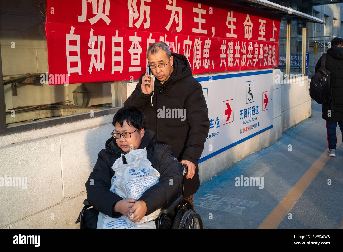 Un père chinois pousse son fils malade en fauteuil roulant tout en parlant sur son téléphone portable dans un hôpital de Pékin, en Chine. 22-Dec-2023 Banque D'Images