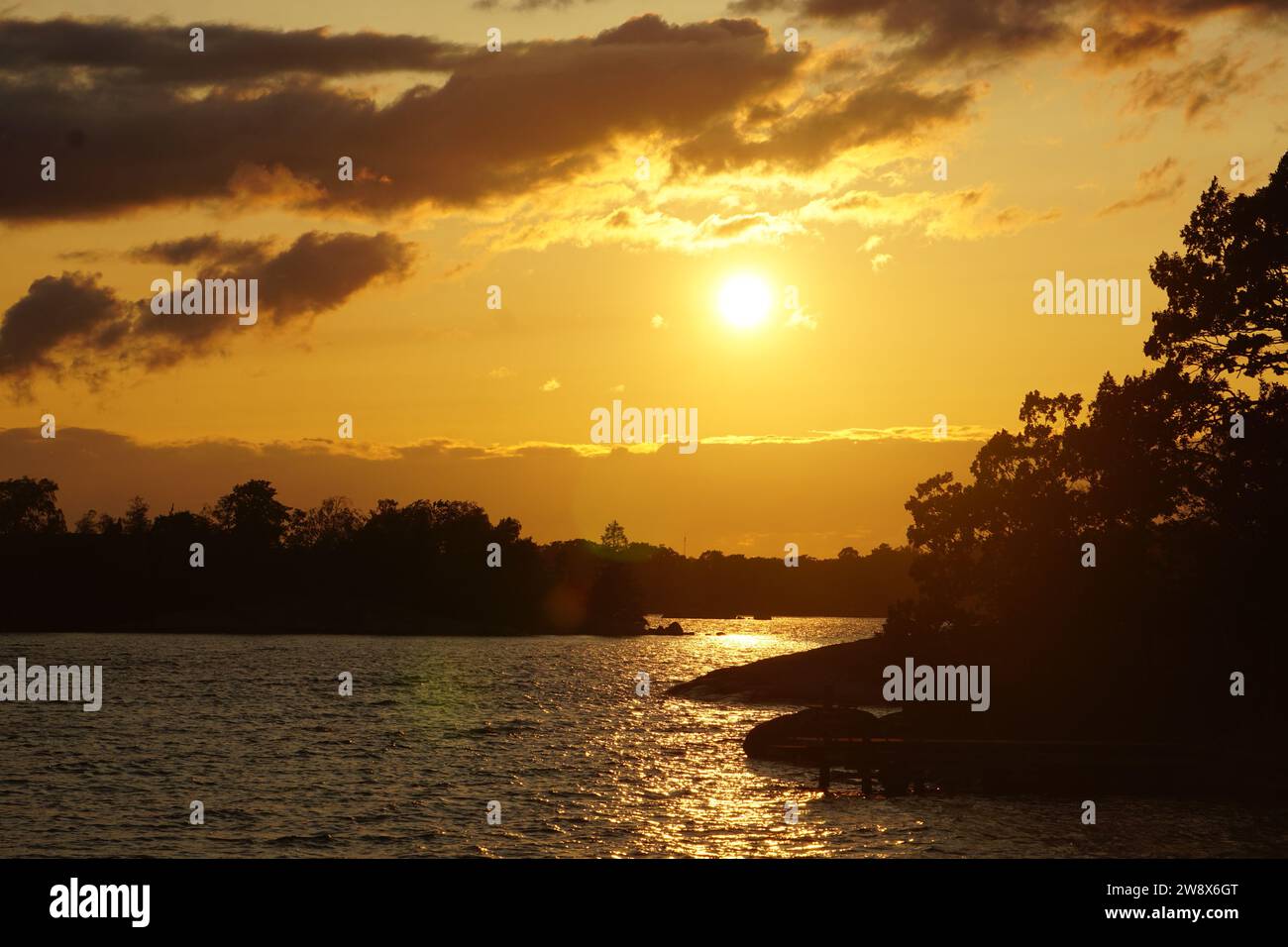 La photo capture un coucher de soleil sur un lac en Suède. Une forêt orne l'horizon, tandis que sur le côté droit, des formations rocheuses sont visibles. Ciel nuageux jaune Banque D'Images