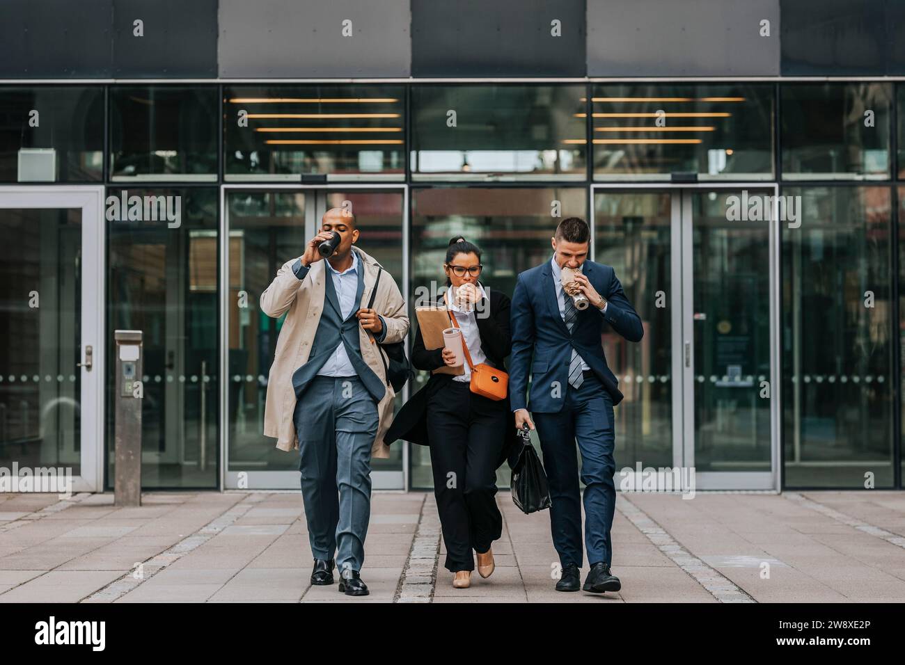 Toute la longueur des collègues hommes et femmes d'affaires ayant de la nourriture tout en marchant devant l'immeuble de bureaux Banque D'Images