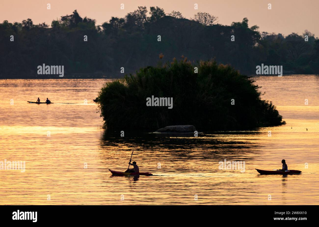 Silhouettes de personnes faisant du kayak à travers les eaux calmes du Mékong, dans l’archipel de si Phan Don, à travers des rayons de lumière dorée réfléchis par la riv Banque D'Images