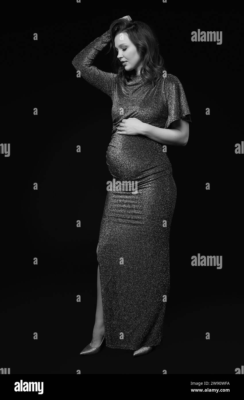 Portrait noir et blanc de jolie femme enceinte sur fond noir de studio. Femme en robe de sequins gris avec les mains près du ventre enceinte. Banque D'Images