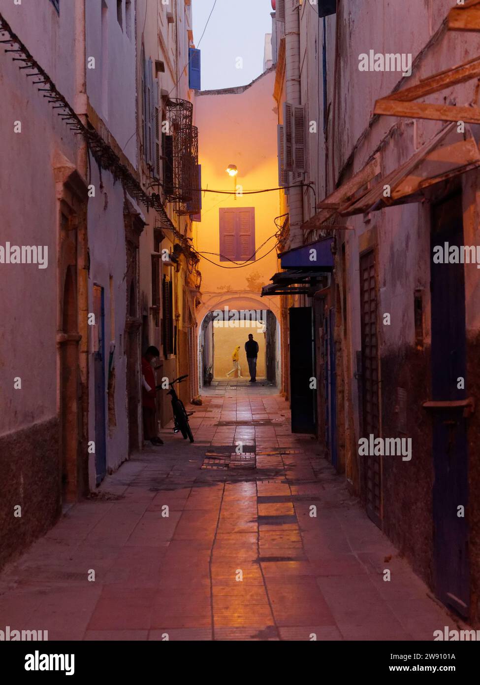 Rue étroite éclairée par un lampadaire la nuit avec un vélo et quelqu'un en silhouette dans la médina à Essaouira, Maroc. 22 décembre 2023 Banque D'Images