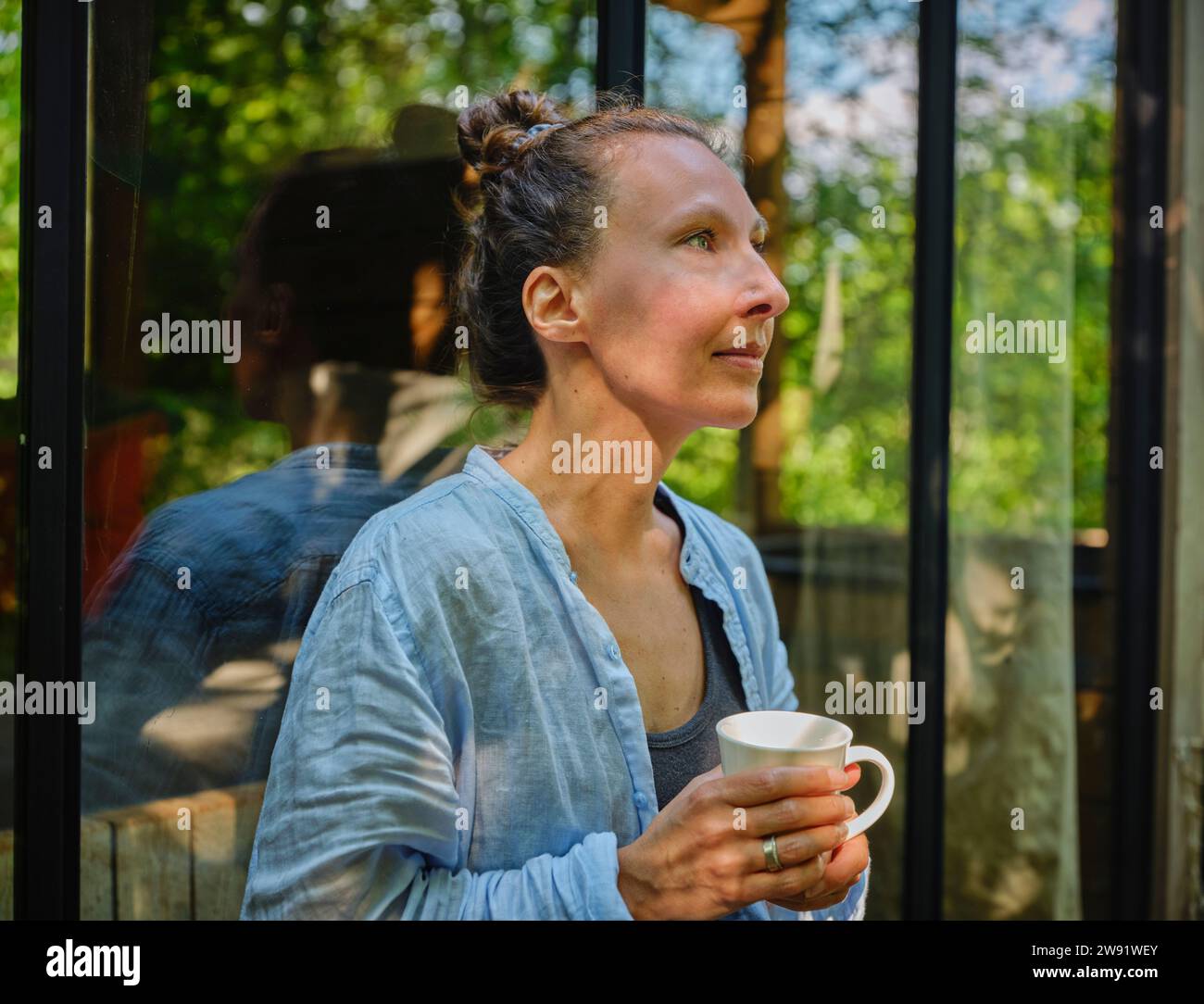 Femme mature tenant une tasse de café et se penchant sur la fenêtre Banque D'Images