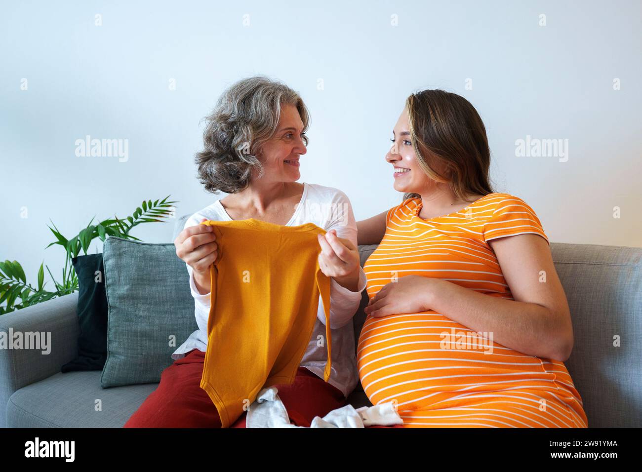 Femme enceinte regardant la mère tenant des vêtements de bébé assis sur le canapé à la maison Banque D'Images