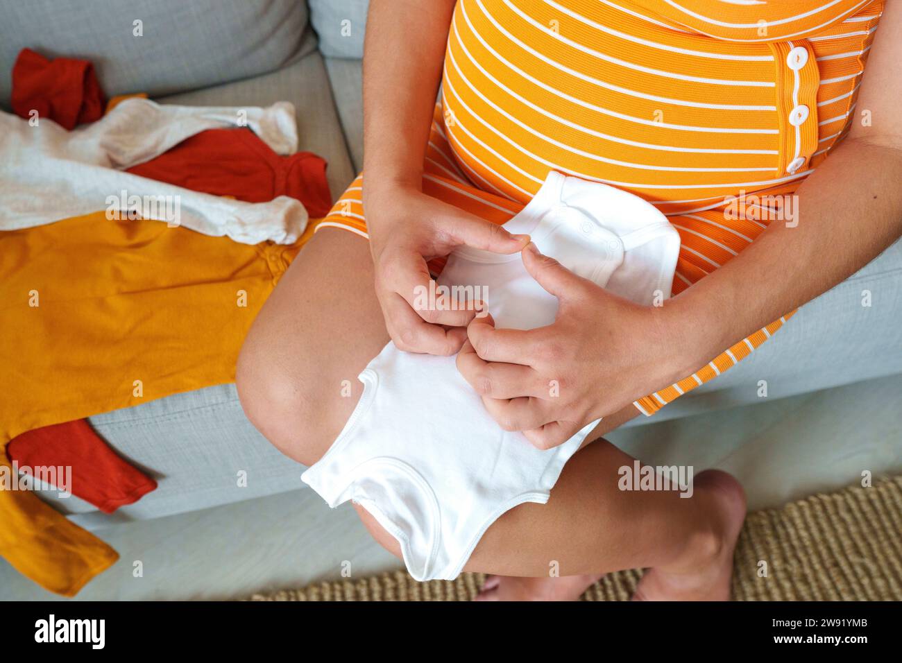 Femme enceinte faisant la forme de coeur sur des vêtements de bébé assis sur le canapé à la maison Banque D'Images