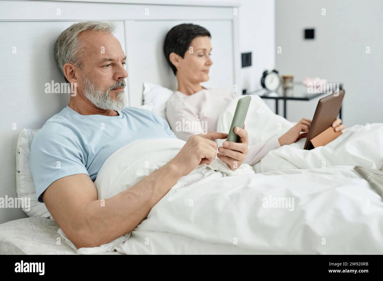 Homme utilisant smartphone tandis que sa femme couchée à côté de lui dans le lit avec tablette numérique le matin Banque D'Images