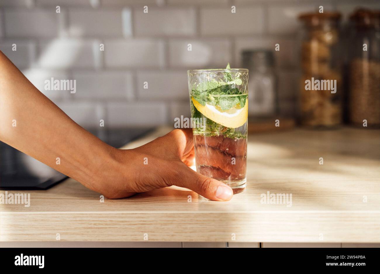 Gros plan de la main féminine tient un mojito citron rafraîchissant sur une table en bois sur une cuisine à la maison. Glace, menthe et citron vert garnissent le cocktail estival. Banque D'Images