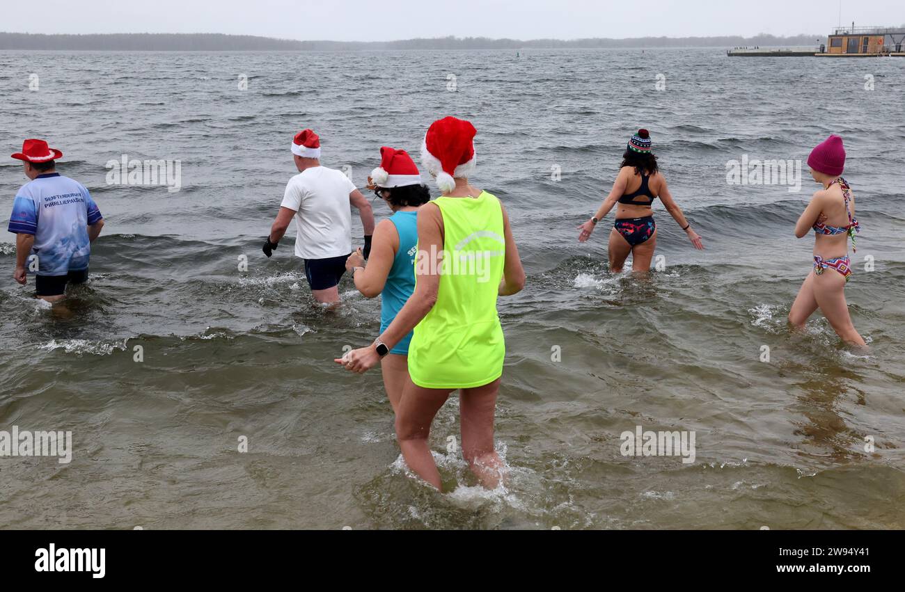 Senftenberg, Allemagne. 24 décembre 2023. Les membres du club de natation d'hiver 'Pirrlliepousen' vont pour leur traditionnelle baignade de Noël dans le lac froid de quatre degrés Senftenberg. La plupart des 25 participants se sont décorés d’un chapeau de Noël. La baignade de Noël a lieu depuis 36 ans - à l'exception de deux ans liés au coronavirus. 'Pirrlliepousen' est un terme nord-allemand pour désigner les glaçons. Crédit : Bernd Wüstneck/dpa/Alamy Live News Banque D'Images