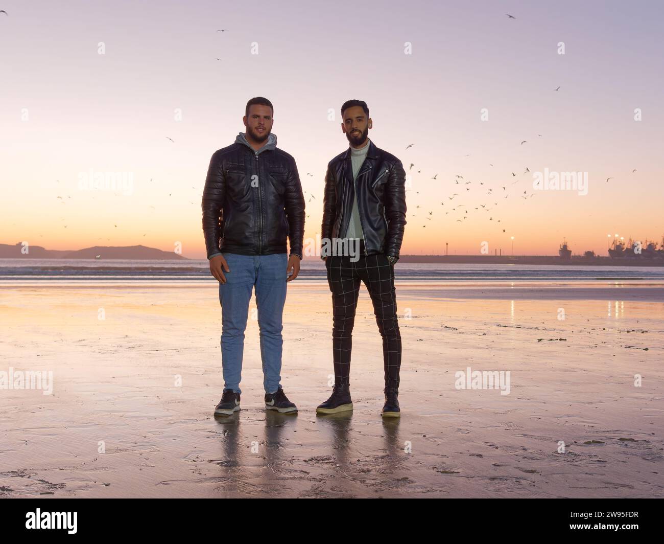 Des amis masculins posent pour un portrait avec les mains dans sa poche, sur une plage de sable au coucher du soleil à Essaouira, Maroc. 23 décembre 2023 Banque D'Images